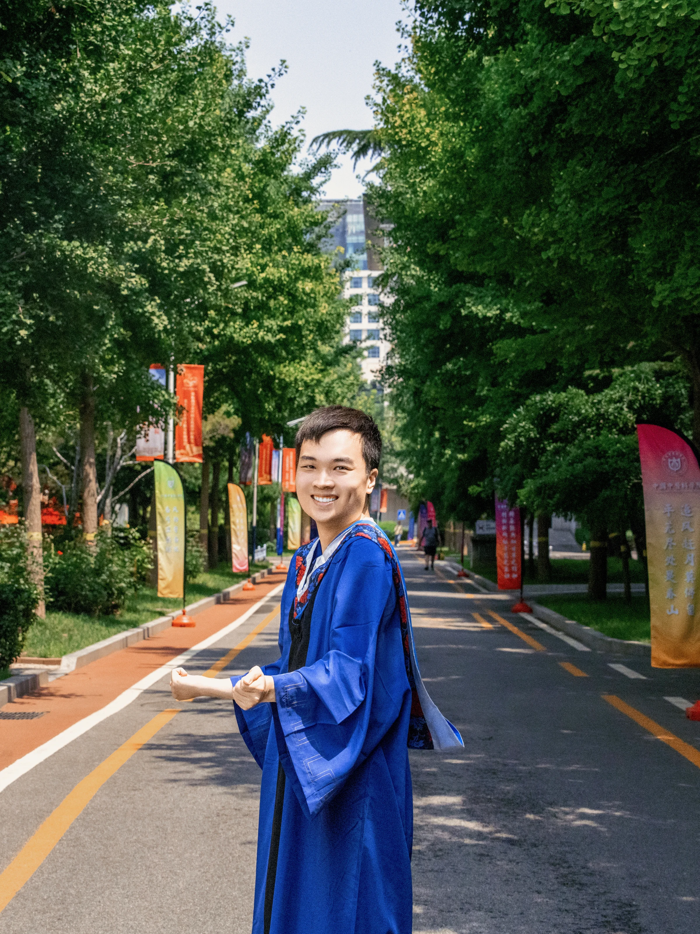 一位身穿蓝色硕士服的年轻男子站在绿树成荫的道路上，面带微笑回望镜头，道路两侧立有彩色道旗。
